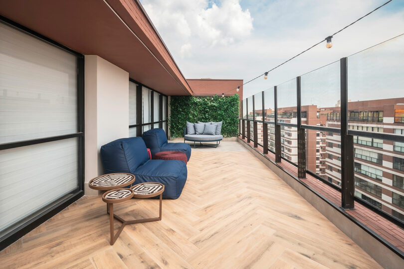 Modern rooftop terrace with wood-pattern floor, blue cushioned seating, geometric tables, string lights, and a green plant wall, overlooking city buildings under a partly cloudy sky.
