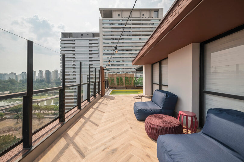 Modern rooftop terrace with glass railings, wooden flooring, blue lounge chairs, small tables, and a view of tall apartment buildings and a park area in the background.