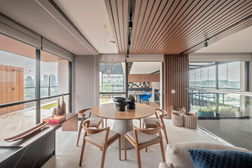 Modern dining area with a round wooden table and four chairs, large windows, wood panel ceiling, neutral tones, and city views in the background.