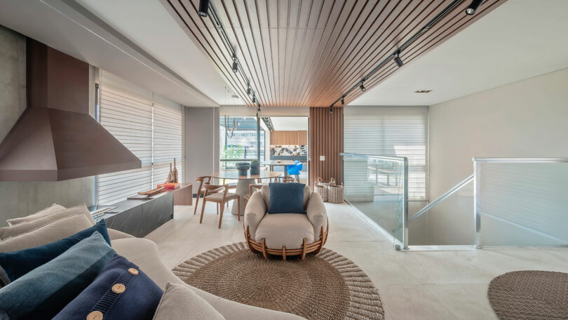 Modern living room with neutral tones, a round woven rug, cushioned armchair, built-in fireplace, dining table, and large windows with blinds; glass railing borders a stairway.