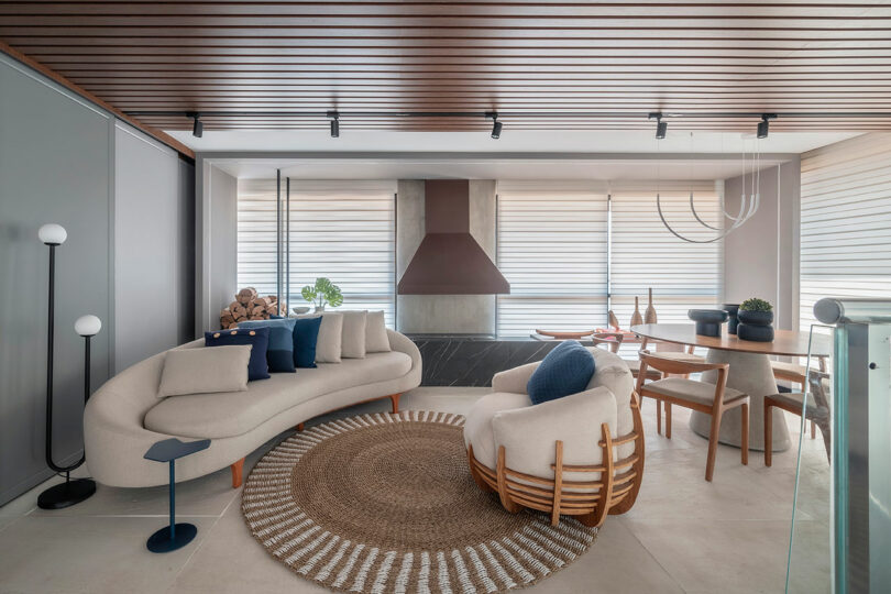 Modern living and dining area with neutral-toned furniture, a round woven rug, a sculptural fireplace, wood accents, and large windows with horizontal blinds.