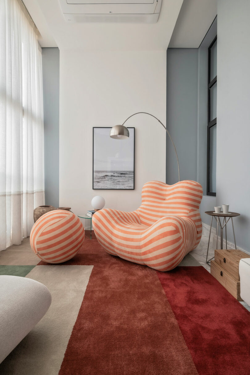 A modern living room with a striped orange armchair and matching ottoman, floor lamp, framed wall art, and a red-and-beige area rug.