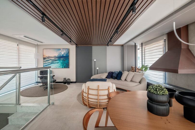 Modern living and dining area with a curved sofa, wooden ceiling slats, round table, indoor plants, and large windows with blinds. A coastal painting hangs on the wall near an armchair.