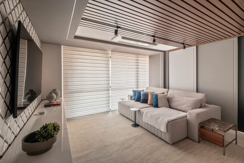 Modern living room with a light gray sectional sofa, small tables, decorative pillows, wall-mounted TV, and large window with blinds, featuring wood ceiling panels and minimalist decor.