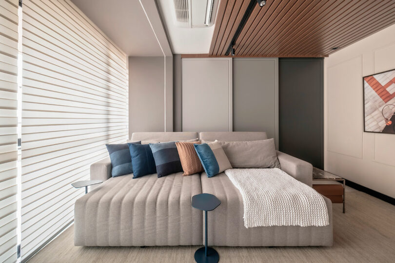 Modern living room with a light gray sectional sofa, assorted blue and brown pillows, a knit throw, small side table, and neutral wall paneling with a framed abstract artwork.