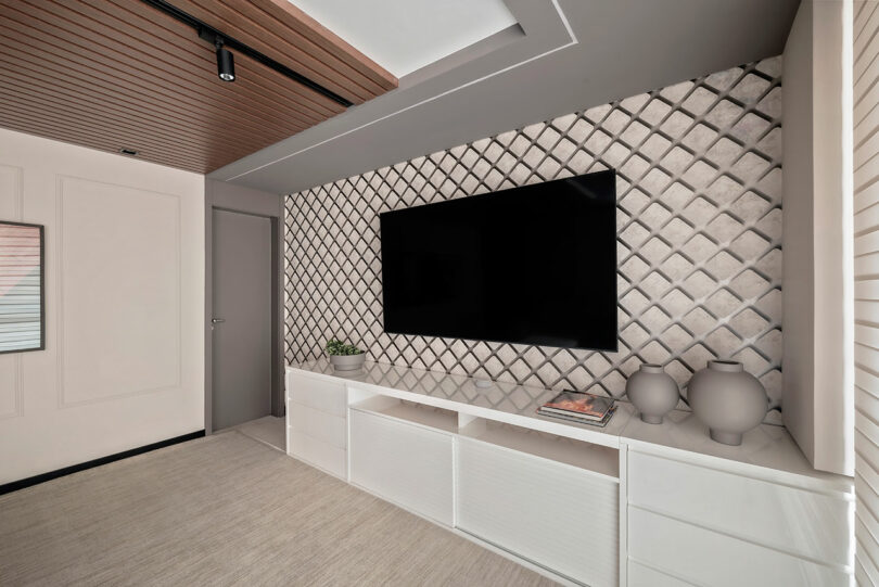 A modern living room features a wall-mounted flat-screen TV, geometric patterned accent wall, white cabinets, and minimalist decor with two vases and a small plant.