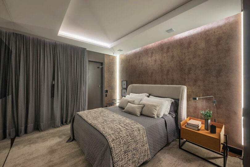 Modern bedroom with a gray upholstered bed, neutral bedding, a wooden nightstand with books and a plant, and soft lighting from a recessed ceiling and wall strip.