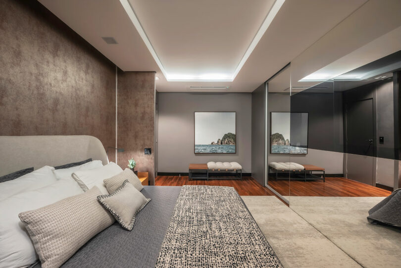 Modern bedroom with neutral tones, a large bed with patterned bedding, wall art of ocean rocks, mirrored closet doors, wooden floors, and recessed ceiling lighting.