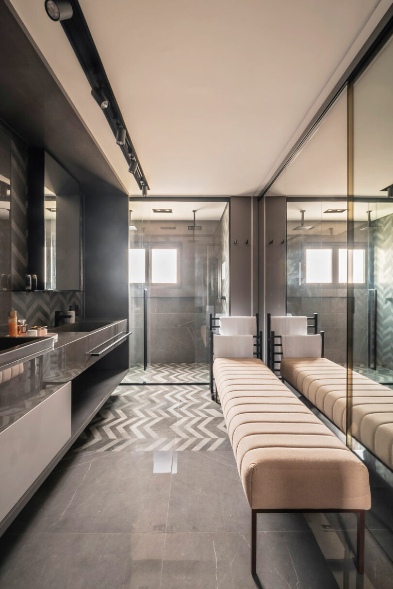 Modern bathroom with chevron-patterned flooring, a long upholstered bench, large mirror, glass shower enclosure, and minimalist sink area.