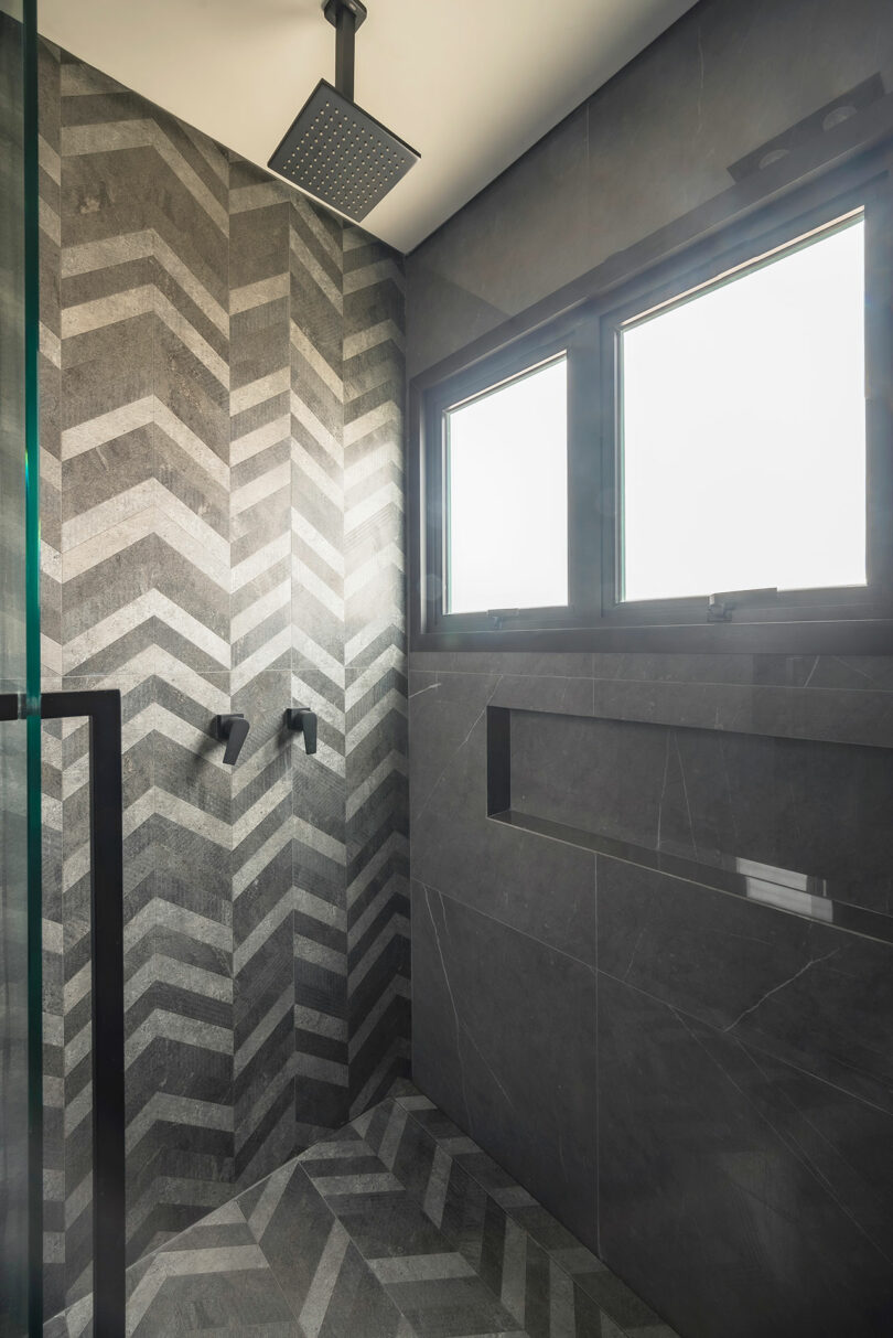 Modern shower with gray herringbone tile pattern, black rainfall showerhead, dual wall controls, built-in shelf, and frosted windows allowing natural light.