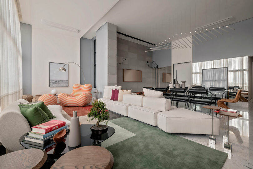 Modern living room with white sectional sofa, striped lounge chair, abstract rugs, glass tables, books, and large windows with sheer curtains, leading to an adjacent dining area.