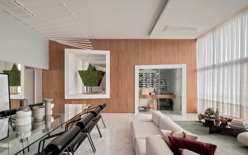 Modern living room with wood-paneled wall, glass dining table, black chairs, white sofa, wine storage display, large window with sheer curtains, and decorative plants.