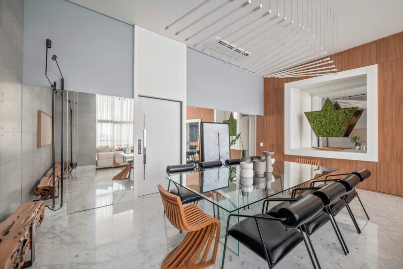 Modern dining room with a glass table, mixed wooden and black chairs, marble floor, wood-paneled walls, large windows, and a decorative green plant feature in the background.