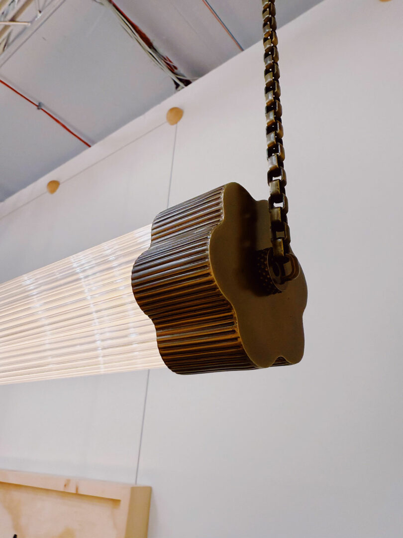 Close-up of a suspended light fixture with a grooved, flower-shaped metal end cap and a metal chain, set against a white ceiling and wall—modern details.