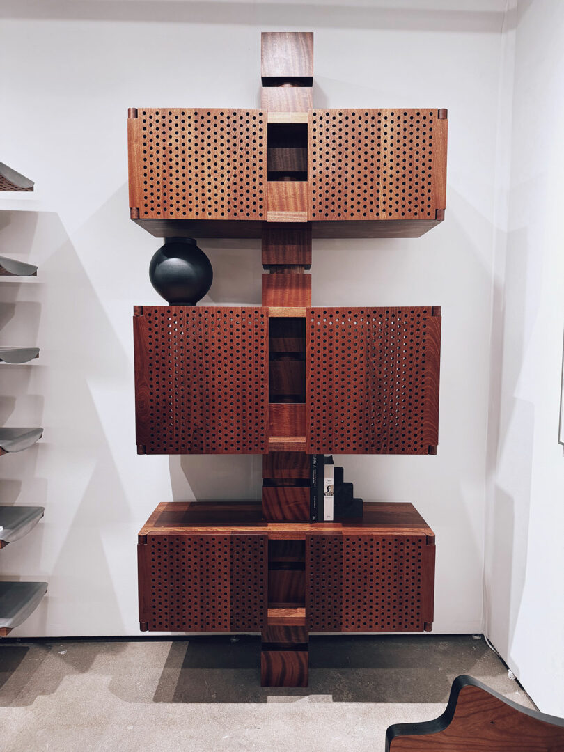 A modern wooden shelving unit featuring perforated cabinet doors, displays a black vase and a few books as it stands elegantly against a white wall.