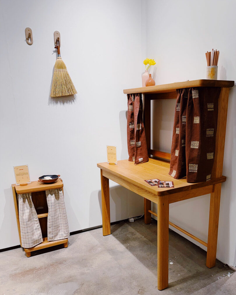 A small wooden desk and side table with patterned curtains, a broom on the wall, and a vase with yellow flowers create a calming minimalist room.