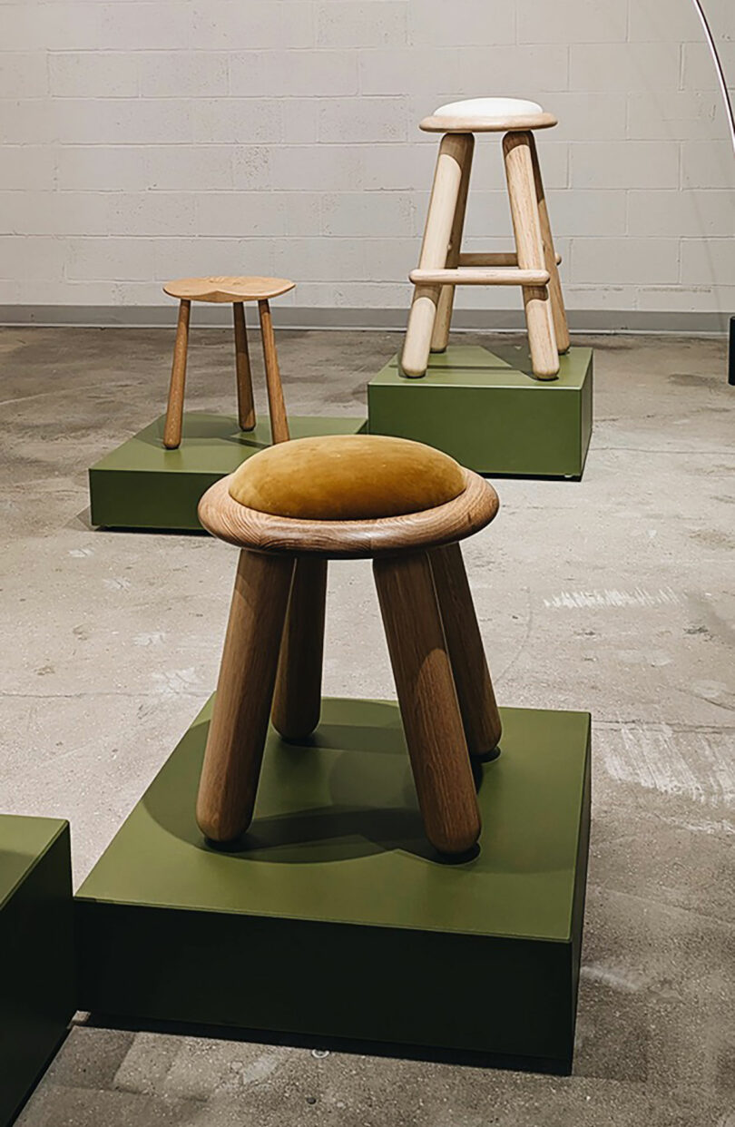 Three wooden stools of varying heights and designs are displayed on green platforms in a minimalist indoor setting, echoing the modern aesthetic envisioned for Shelter 2025, with a concrete floor and white brick wall.
