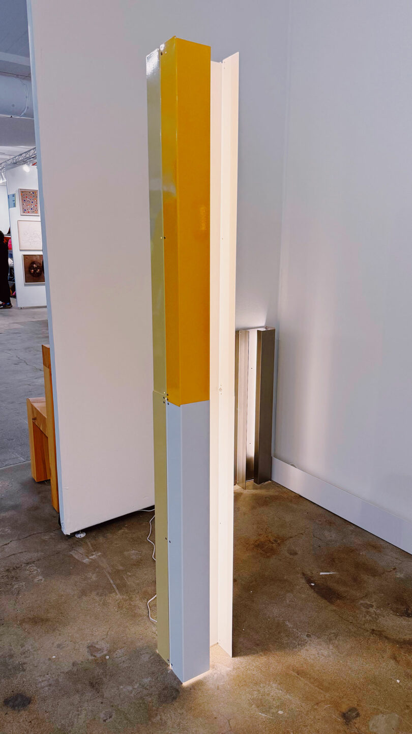 A tall, rectangular, geometric sculpture features vertical panels in yellow, white, and light gray standing on a concrete floor in an art gallery.