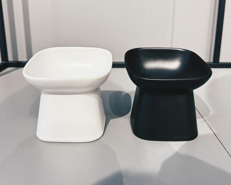 Two modern, square-shaped bowls with wide rims—one white and one black—are placed side by side on a gray surface, showcasing a sleek aesthetic perfect for contemporary spaces like those envisioned in Shelter 2025.
