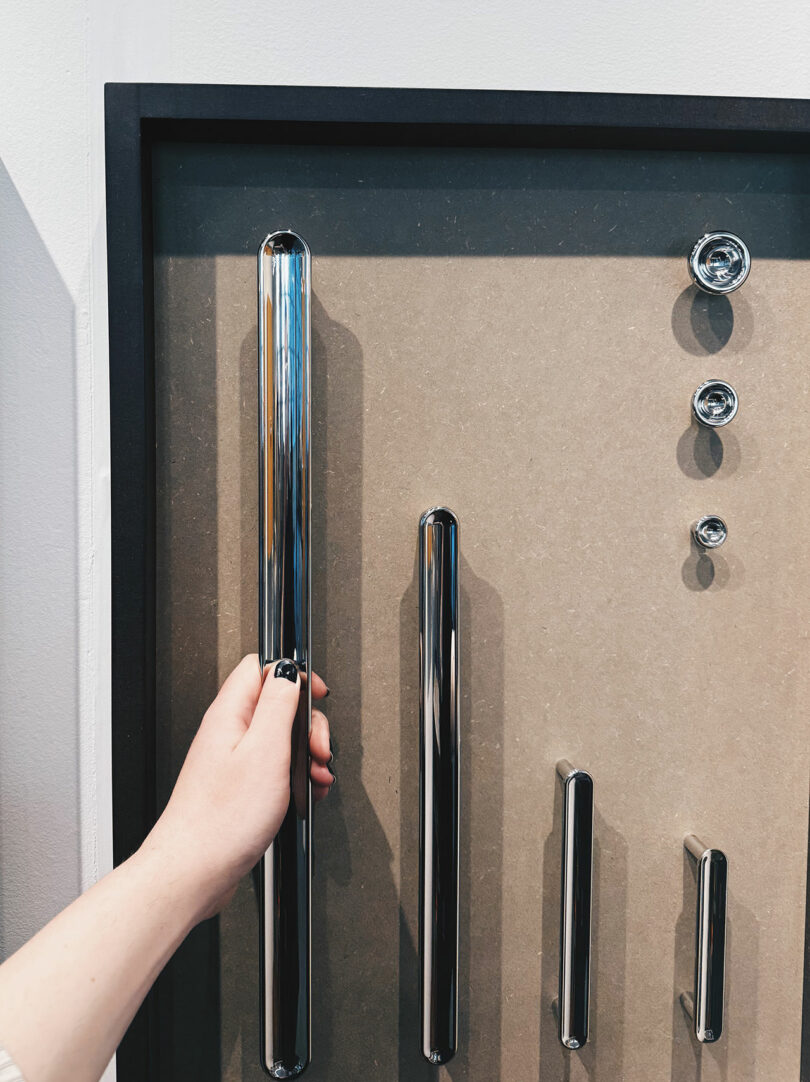 A hand grasps a long, metallic door handle on a display board featuring several other handles and round fixtures mounted on it.