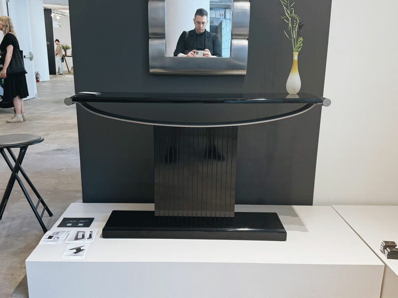 A modern black console table with a curved metal accent is displayed on a white platform, with a vase and a mirror reflecting a person above it.