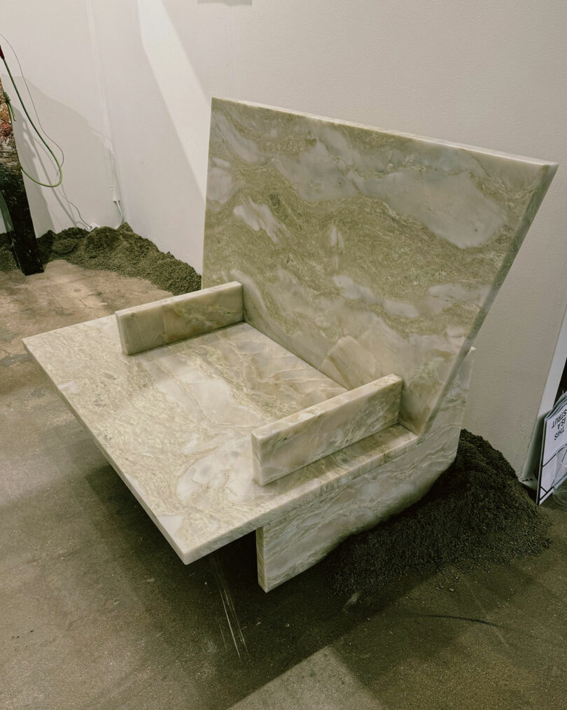 A chair made of polished stone with a large, angled backrest and rectangular armrests, is displayed on a concrete floor with dark sand surrounding its base.