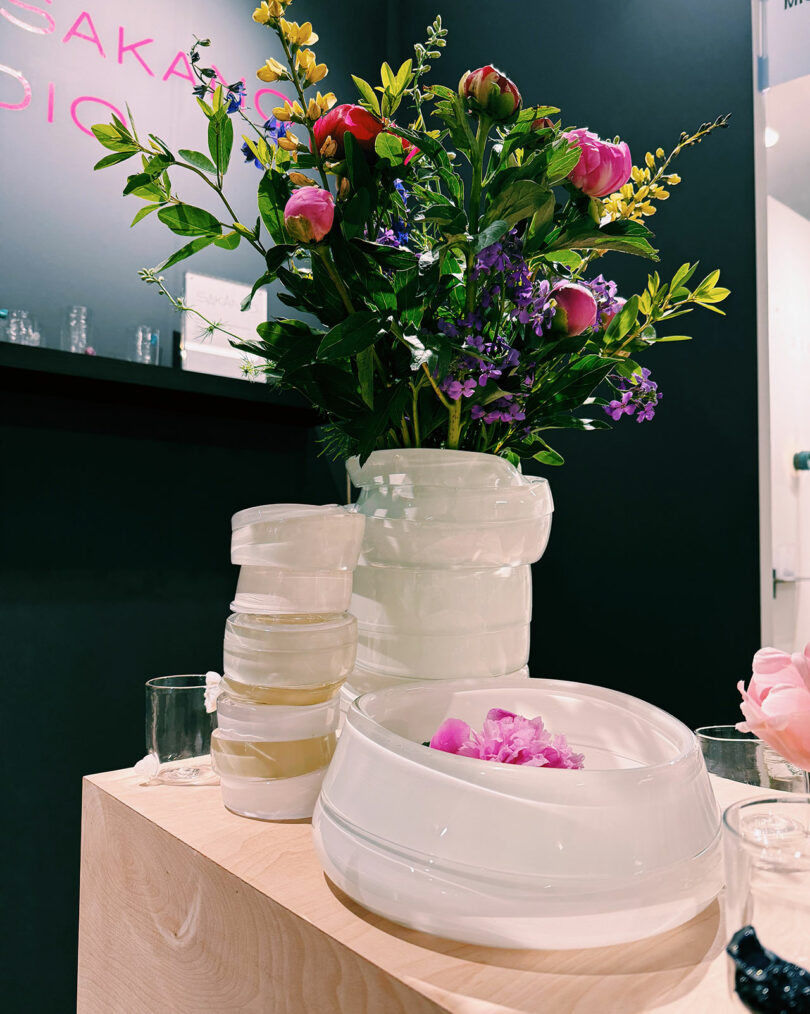 A white vase with colorful flowers, several white bowls, and glass containers are displayed on a wooden surface against a dark background.