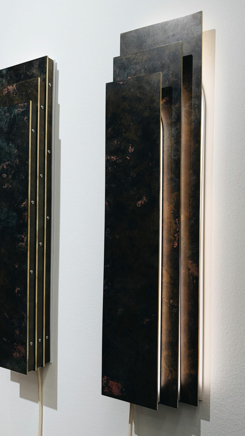 Close-up of a modern, rectangular wall-mounted sconce featuring layered metal panels and soft backlighting, attached to a white wall.