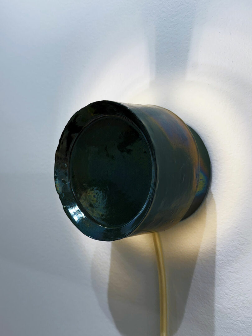 A metallic, bowl-shaped object is mounted on a white wall, with a cord extending downward from its base, casting a soft shadow behind it—a subtle detail in the minimalist design of Shelter 2025.
