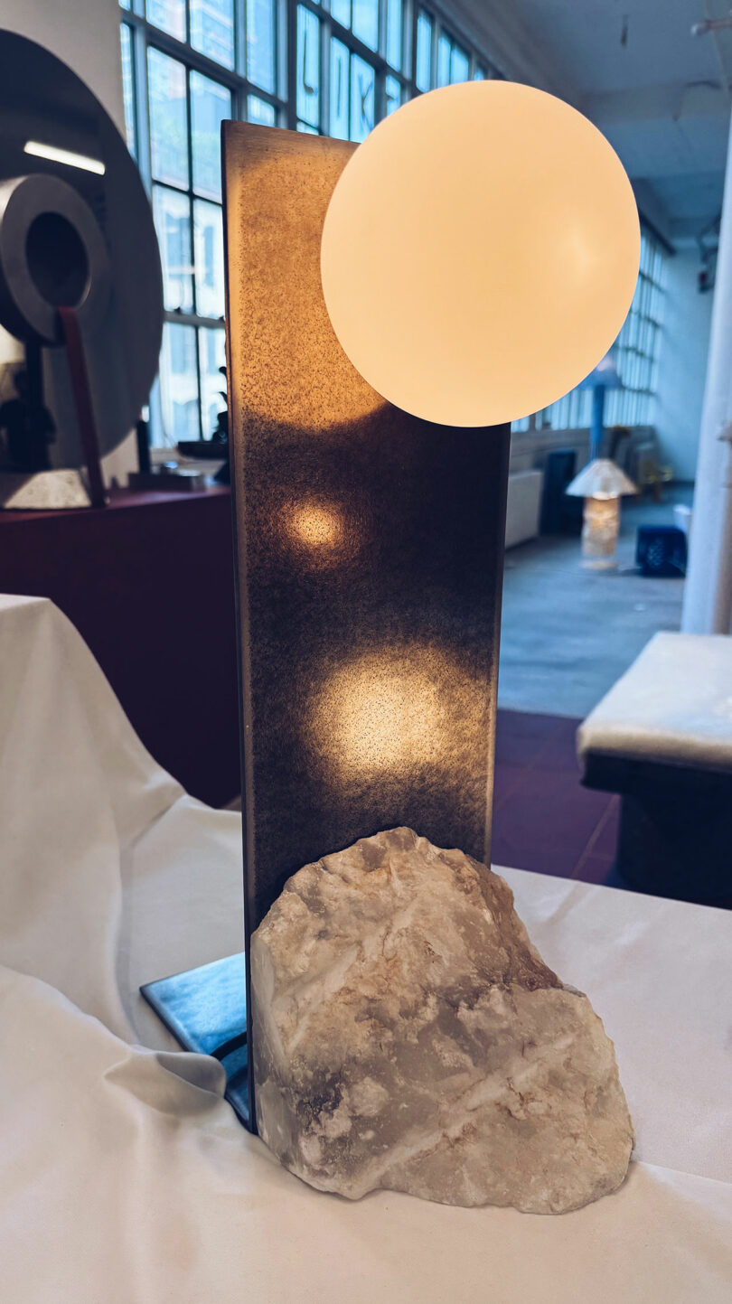 A modern table lamp with a round white bulb, a rectangular metallic stand, and a rough stone base, set on a draped white surface in a spacious interior.