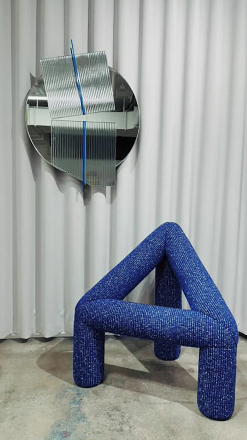 A geometric blue chair with three tubular legs sits in front of white curtains, beneath a round abstract wall sculpture with metallic and blue accents.