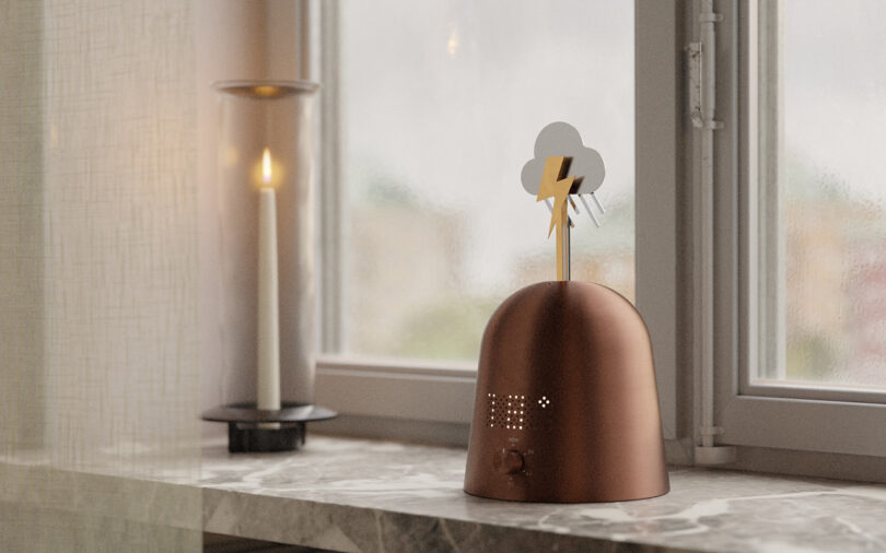 A small brown weather device with a cloud and lightning bolt on top sits on a marble windowsill next to a lit candle in a glass holder.