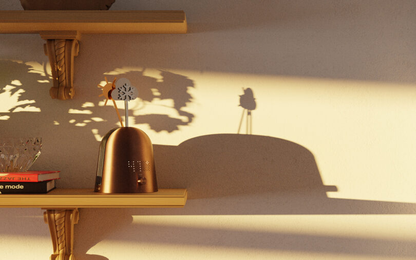 A small metallic device, books, and a glass object are arranged on a wooden wall shelf, casting distinct shadows on a sunlit cream-colored wall.
