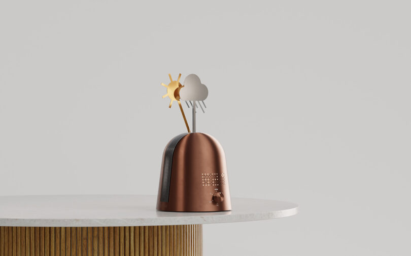 A metallic device with sun, cloud, and raindrop symbols on top sits on a white round table against a plain gray background.