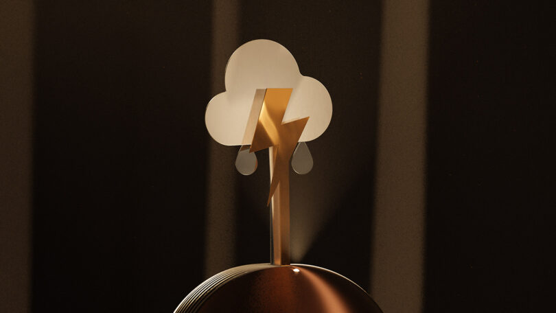 A metallic sculpture of a cloud with raindrops and a lightning bolt stands on a round, reflective base against a dark background.