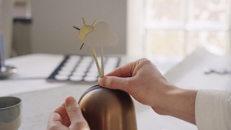 A person holds a metallic object and places a cutout of a sun and cloud on top of it. A bowl and papers are visible on the table in the background.