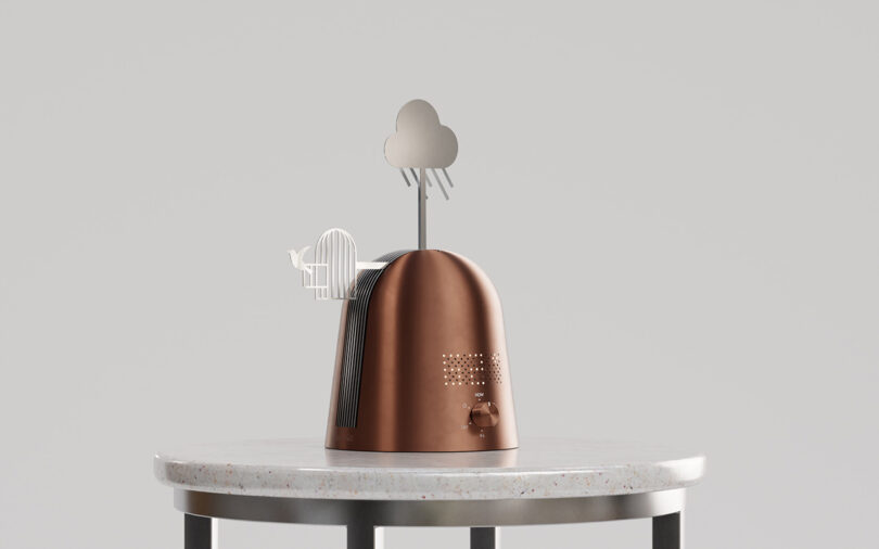A metallic brown device with decorative metal cutouts of a cloud, tree, and bird in a cage, placed on a round white table against a plain background.