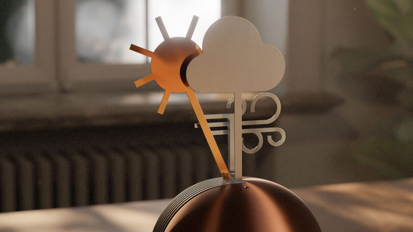 A decorative metallic sculpture of a sun, cloud, and wind on a rounded base sits on a table in front of a window.