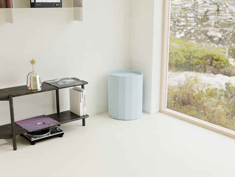 Minimalist corner of a room with a small black shelving unit, a stack of magazines, a decorative object, and a light blue cylindrical bin next to a large window overlooking greenery and rocks.