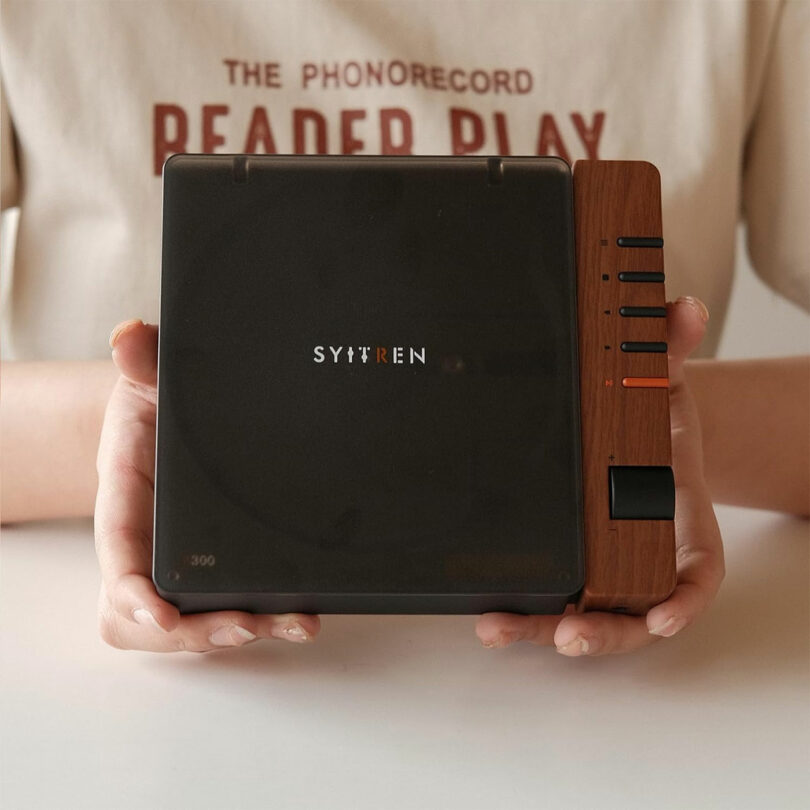 A person holds a compact, square-shaped Syitren R300 audio device with wood accents and black control buttons, against a plain background.