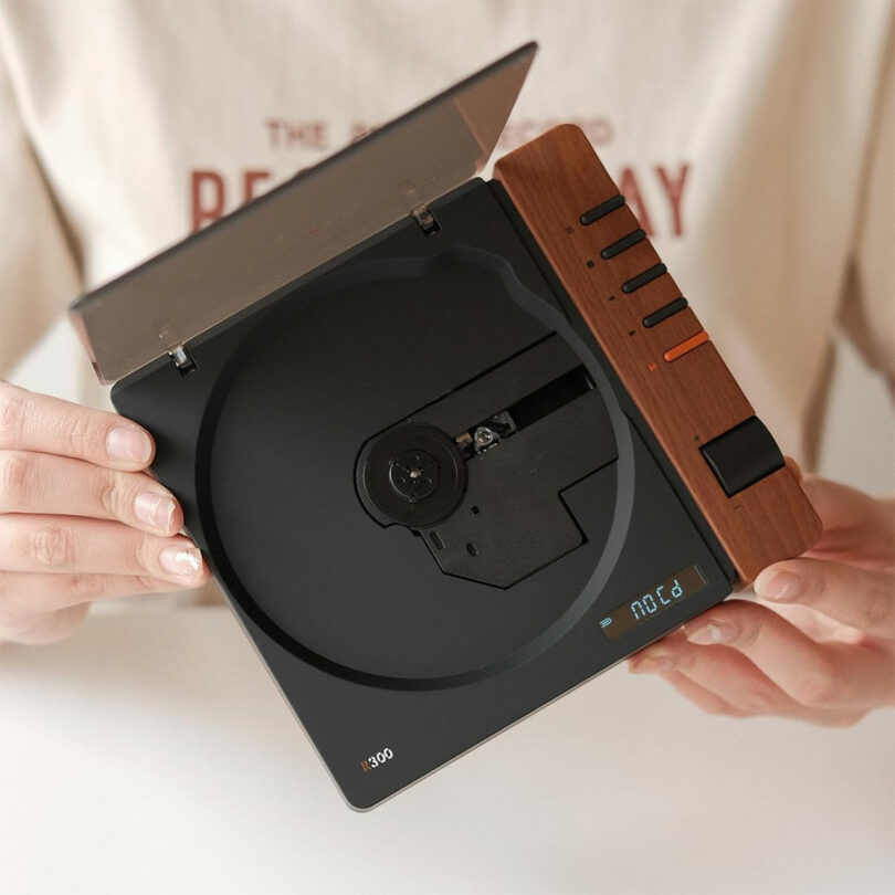 A person holds an open Syitren R300, a square-shaped CD player with a wood finish and digital display, revealing the CD compartment and control buttons.
