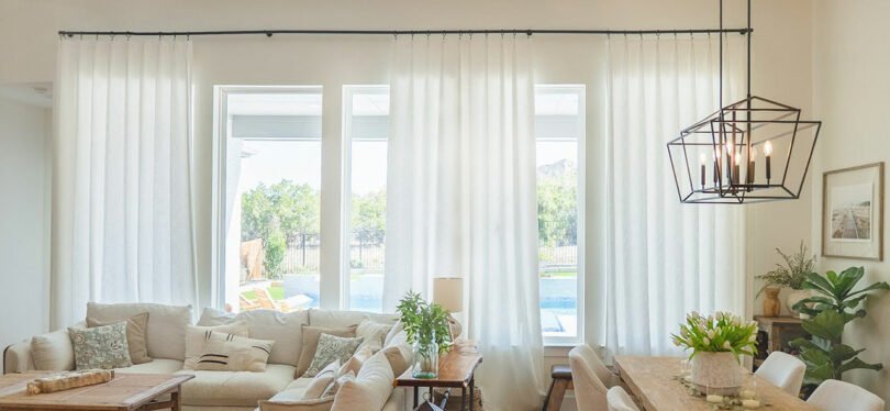 Bright living and dining room with white TWOPAGES curtains, large windows, beige sofas, a wooden table, and modern chandelier, overlooking a garden and pool.