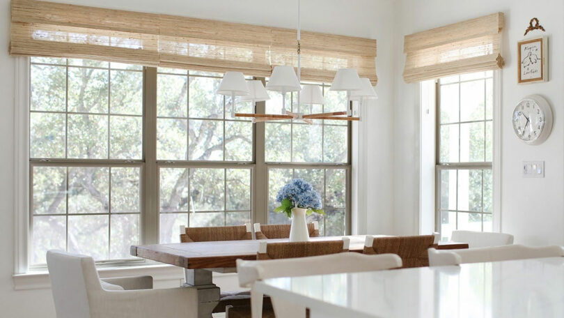 A dining area with a wooden table, white chairs, large windows featuring TWOPAGES bamboo shades, a white pendant light, and a vase of blue flowers on the table.