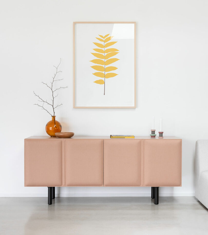 A Tacito Sideboard in a peachy hue stands against a white wall, topped with a vase, bowl, two candlesticks, and a book; a framed yellow leaf print hangs above.