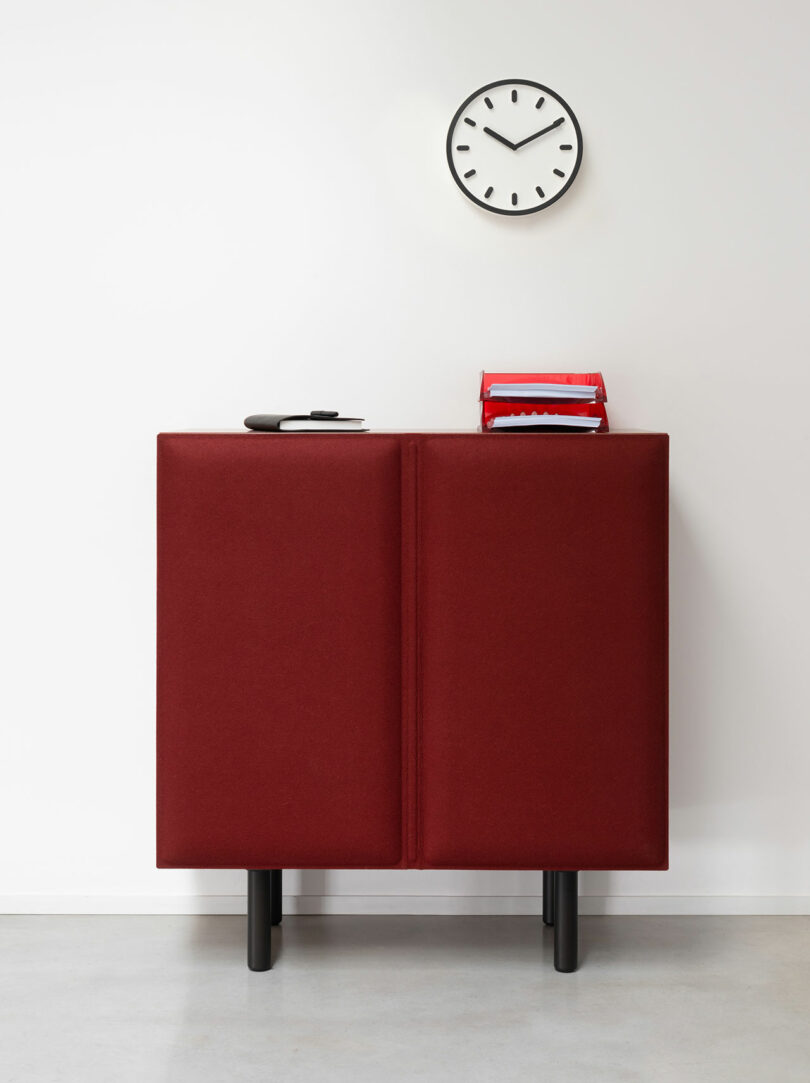 A red Tacito Sideboard with black legs stands against a white wall, topped with stacked red binders and a closed notebook, below a round wall clock showing 11:10.