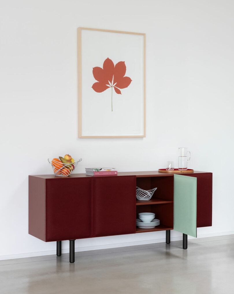 The Tacito Sideboard features burgundy and mint doors for stylish dish storage, topped with fruit, books, and a water carafe. Above, a red flower print accents the white wall for a modern look.