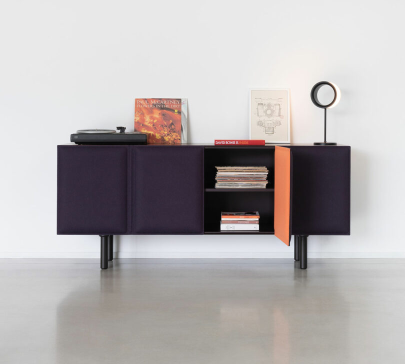 The Tacito Sideboard features dark purple doors, with one orange door open to display books, a turntable, vinyl records, framed art, and a circular lamp on top.