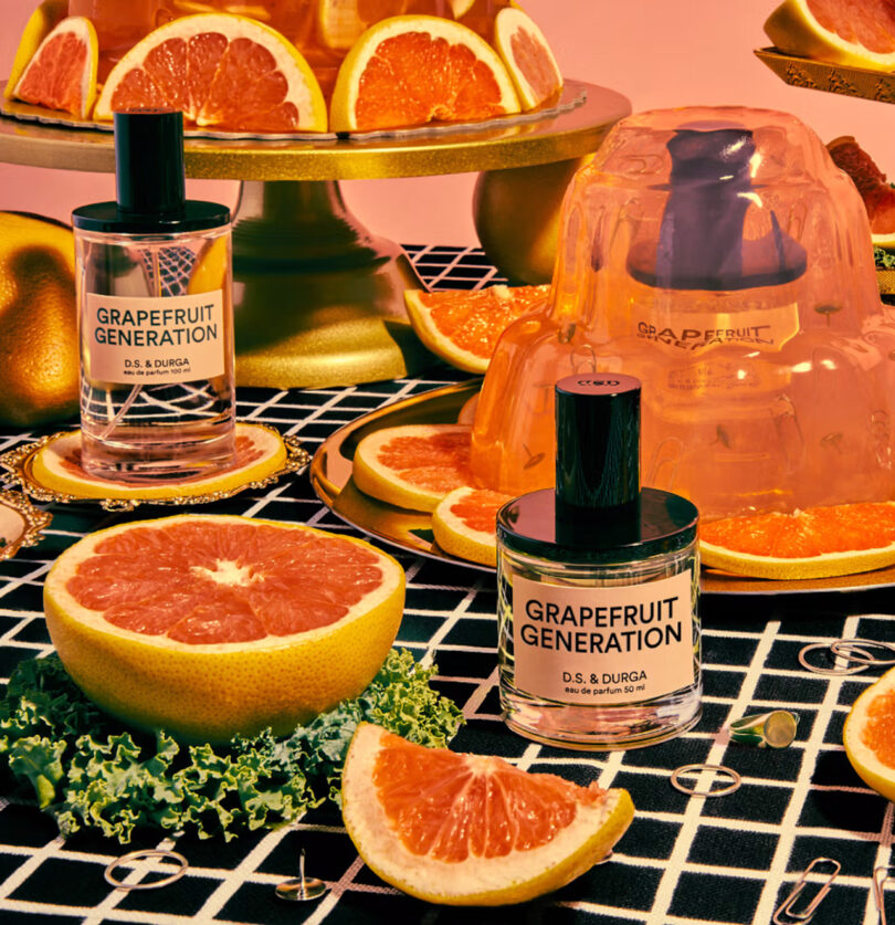 Bottles of "Grapefruit Generation" perfume sit among halved grapefruits, jelly, and leafy greens on a patterned tablecloth with a pink backdrop.