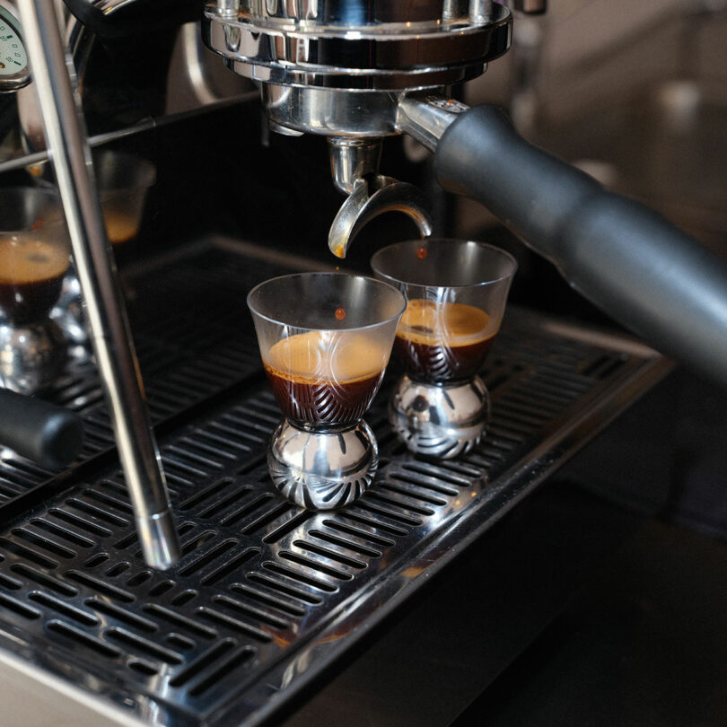 Two glasses from the Artisans Series are placed under an espresso machine as fresh espresso is being dispensed, showing crema on top of the coffee.