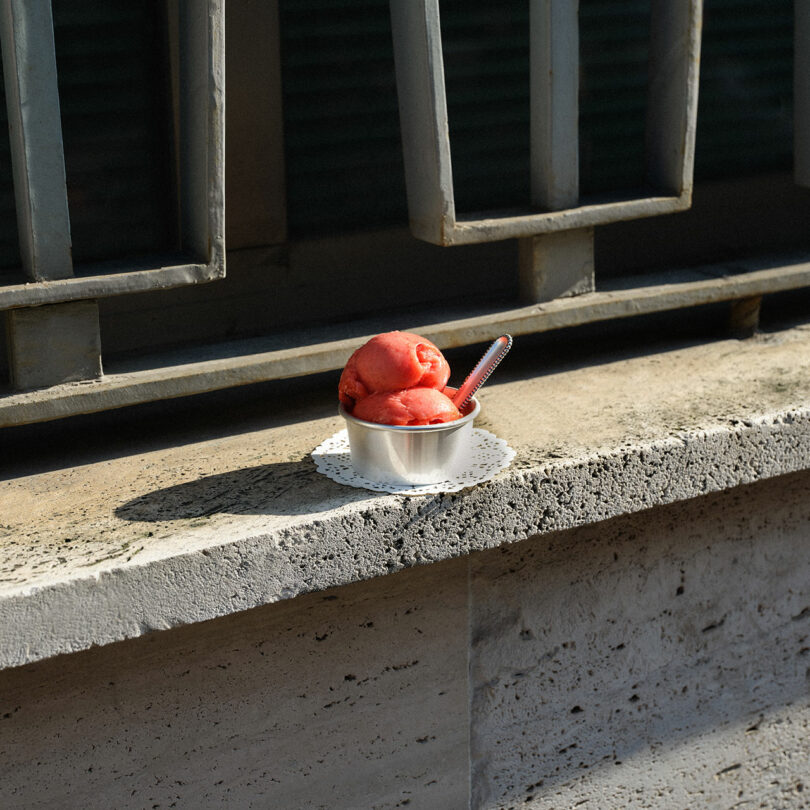 An Artisans Series metal bowl containing red sorbet and a spoon rests on a lace doily placed on a concrete ledge, with metal railings in the background.